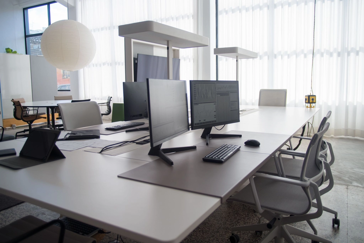Keep clean - Reinigungs - Ein minimalistisches, modernes Großraumbüro zeichnet sich durch weiße Schreibtische mit Computermonitoren, Tastaturen und ergonomischen grauen Stühlen aus, die unter hellem, natürlichem Licht stehen, das durch dünne Vorhänge fällt.